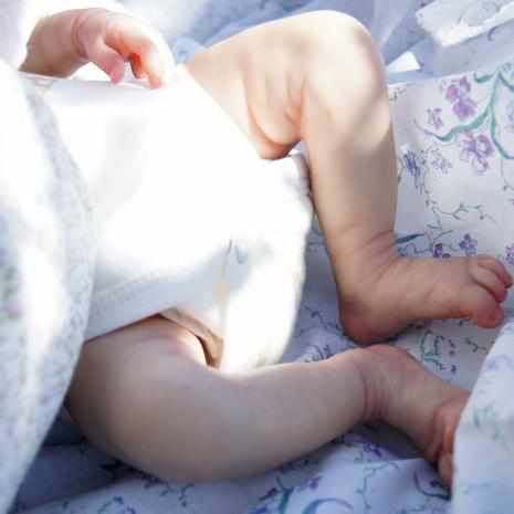 Close-up of a reborn baby girl's legs in white clothing, lying on a floral patterned and white blanket in natural light.