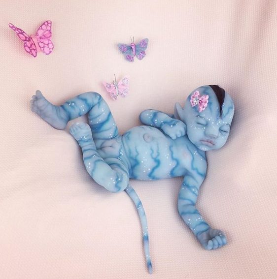 A sleeping baby painted blue with fantasy elements, including feline ears and a tail, surrounded by pink butterfly props.