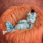 A baby painted as a blue alien sleeps on an orange, furry blanket, adorned with a floral wreath on its head.