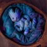 A baby painted in blue and purple hues with white star-like dots, resembling a galaxy, sleeps in a cozy nest of blue fabric.