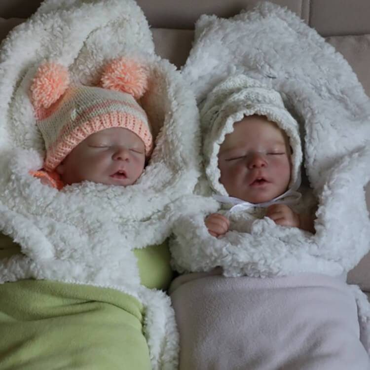 Two swaddled reborn babies sleeping: one in a green blanket with a pink hat, the other in a pink blanket with a white hat.
