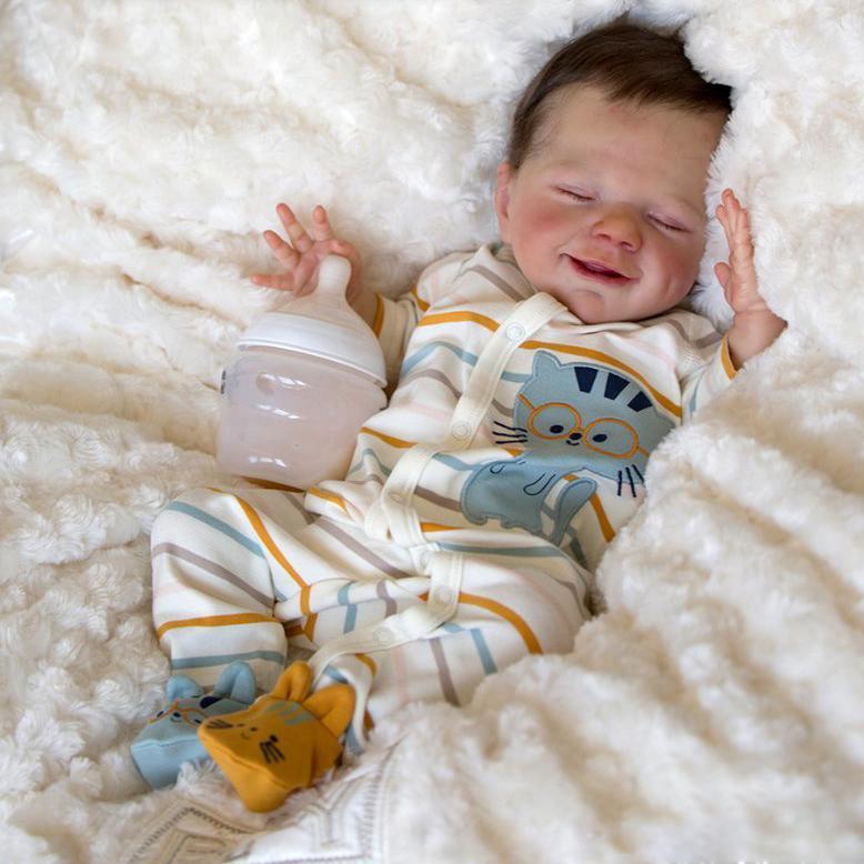 Smiling reborn baby boy in striped pajamas holding a bottle, lying on a soft white blanket.