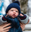 A realistic doll dressed as a baby boy in a navy blue beanie and jacket is held by someone with manicured nails, with a blurred outdoor background.
