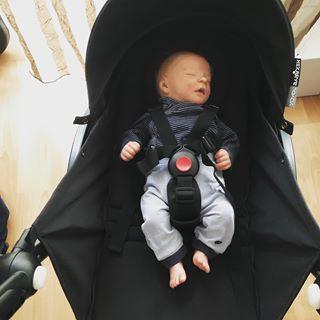 A reborn baby girl wearing a striped shirt and light pants sleeps in a black stroller, secured with a harness.