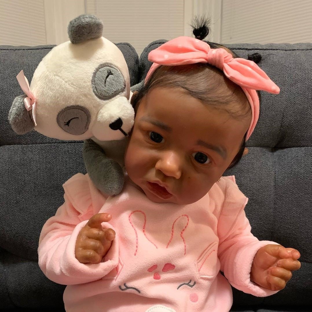 Baby in pink outfit with a matching bow headband, sitting on a couch and holding a stuffed animal panda toy on the shoulder, looking just like one of those realistic dolls.