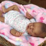 A sleeping reborn baby boy in a floral onesie rests on a pink patterned blanket in a woven basket.