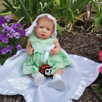 A realistic reborn baby girl in a green dress and white bonnet sits on a blanket surrounded by flowers, holding a ladybug toy.