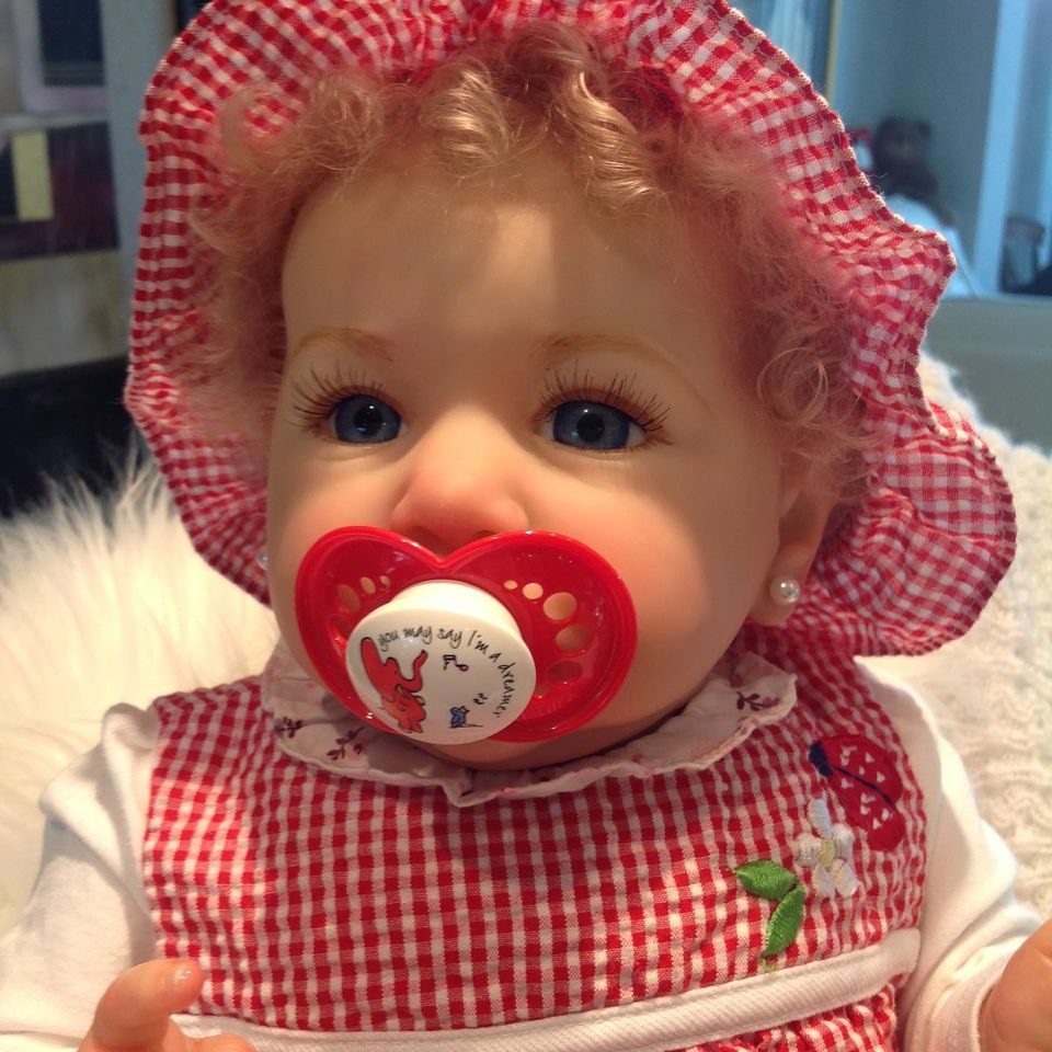 A reborn baby girl with curly hair, blue eyes, and a red pacifier wears a red gingham bonnet and matching dress adorned with a flower.