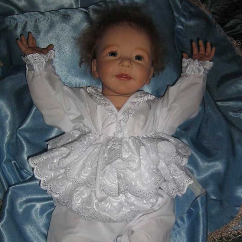 A lifelike reborn baby boy wearing a frilly white outfit lies on a blue satin blanket with arms stretched upward, capturing the essence of innocence and comfort.