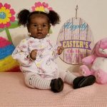 A reborn baby girl with curly hair, wearing a bunny outfit and headband, sits near Easter decor and stuffed toys.