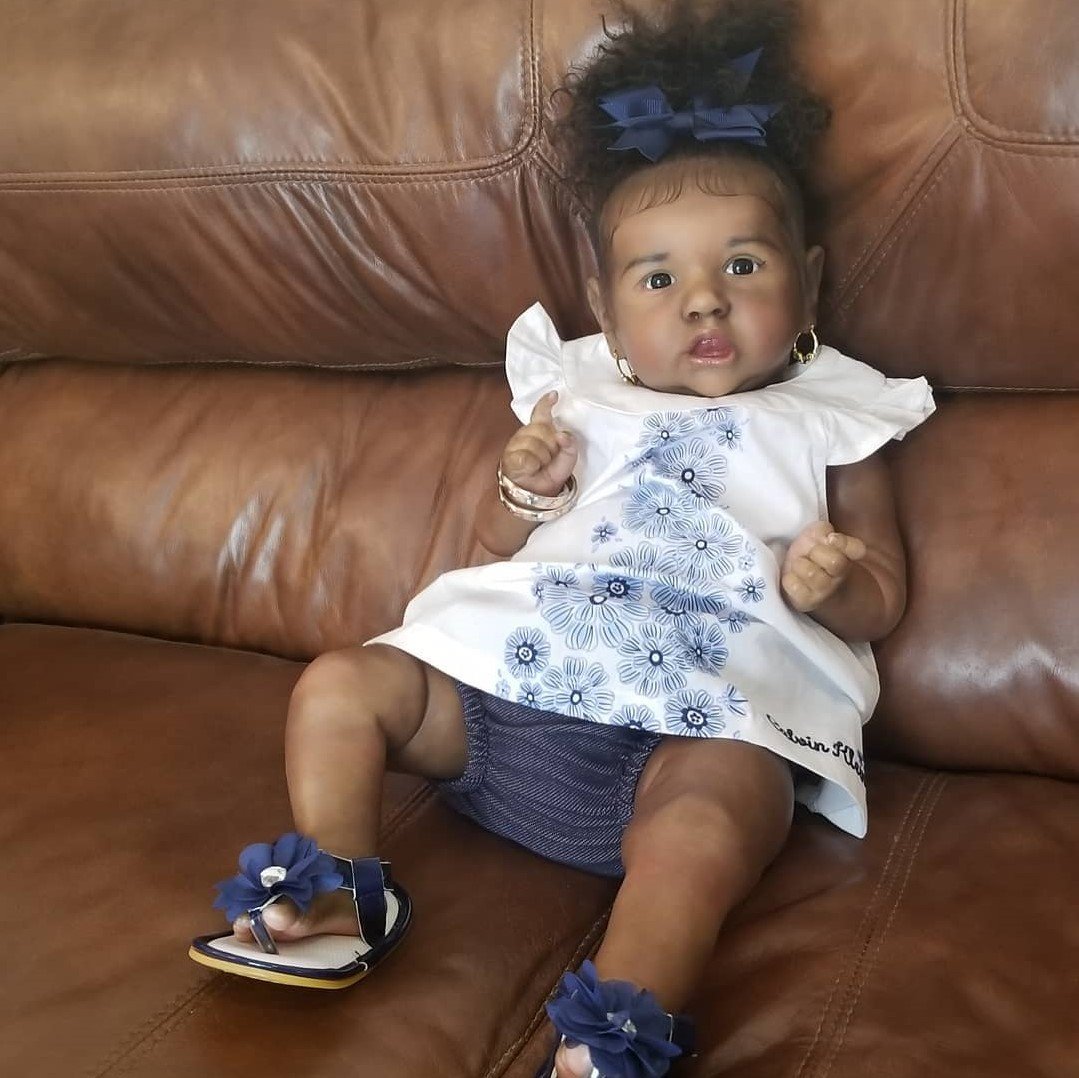 A reborn doll dressed in a blue and white outfit and sandals, sitting on a brown leather couch.